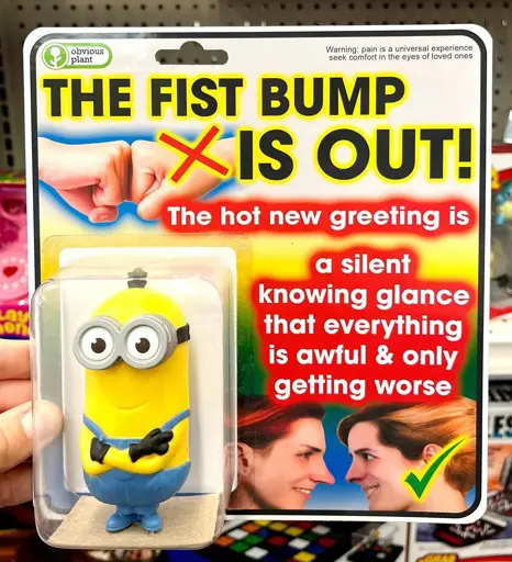 Fake product created by Obvious Plant: a packaged Minion figure from Despicable Me. There is a crossed out fist bump on the package, as well as two people's heads staring intently at each other. The label says "The fist bump is out! The hot new greeting is a silent knowing glance that everything is awful & only getting worse". Warning text in the top right says "Pain is a universal experience - seek comfort in the eyes of loved ones."